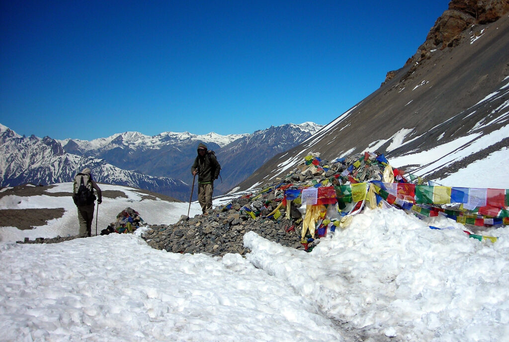 Trekkers crossing Thorong La Pass on the Annapurna Circuit with prayer flags and mountain peaks.