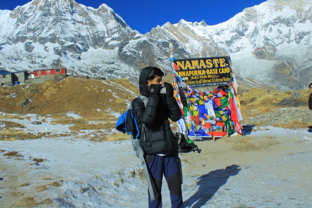 Trekker standing in front of the welcome board at Annapurna Base Camp with snow-capped peaks in the background.