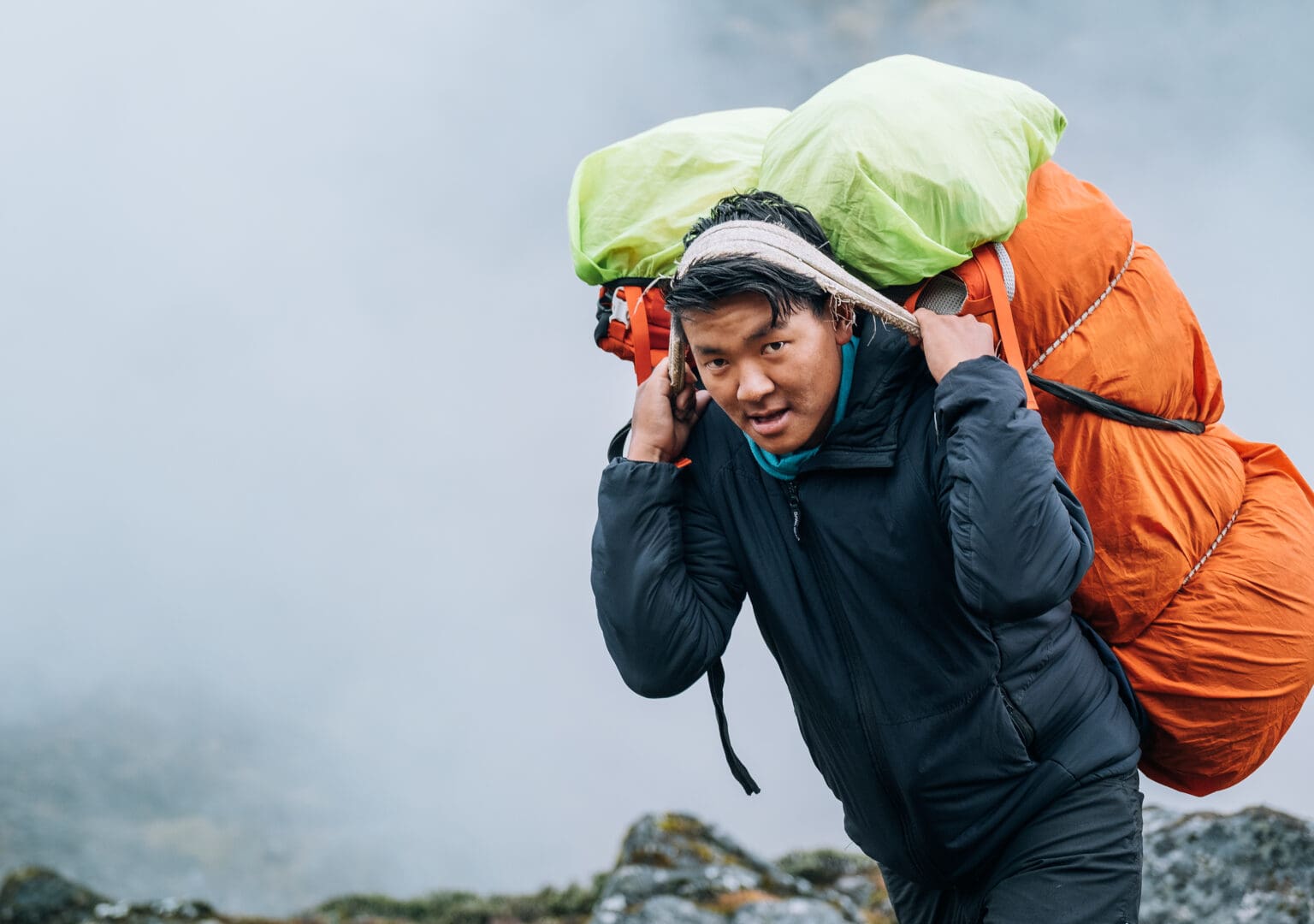 A determined porter navigating the rugged terrain of the Himalayas, balancing a heavy load on his back as he journeys through the scenic landscape.