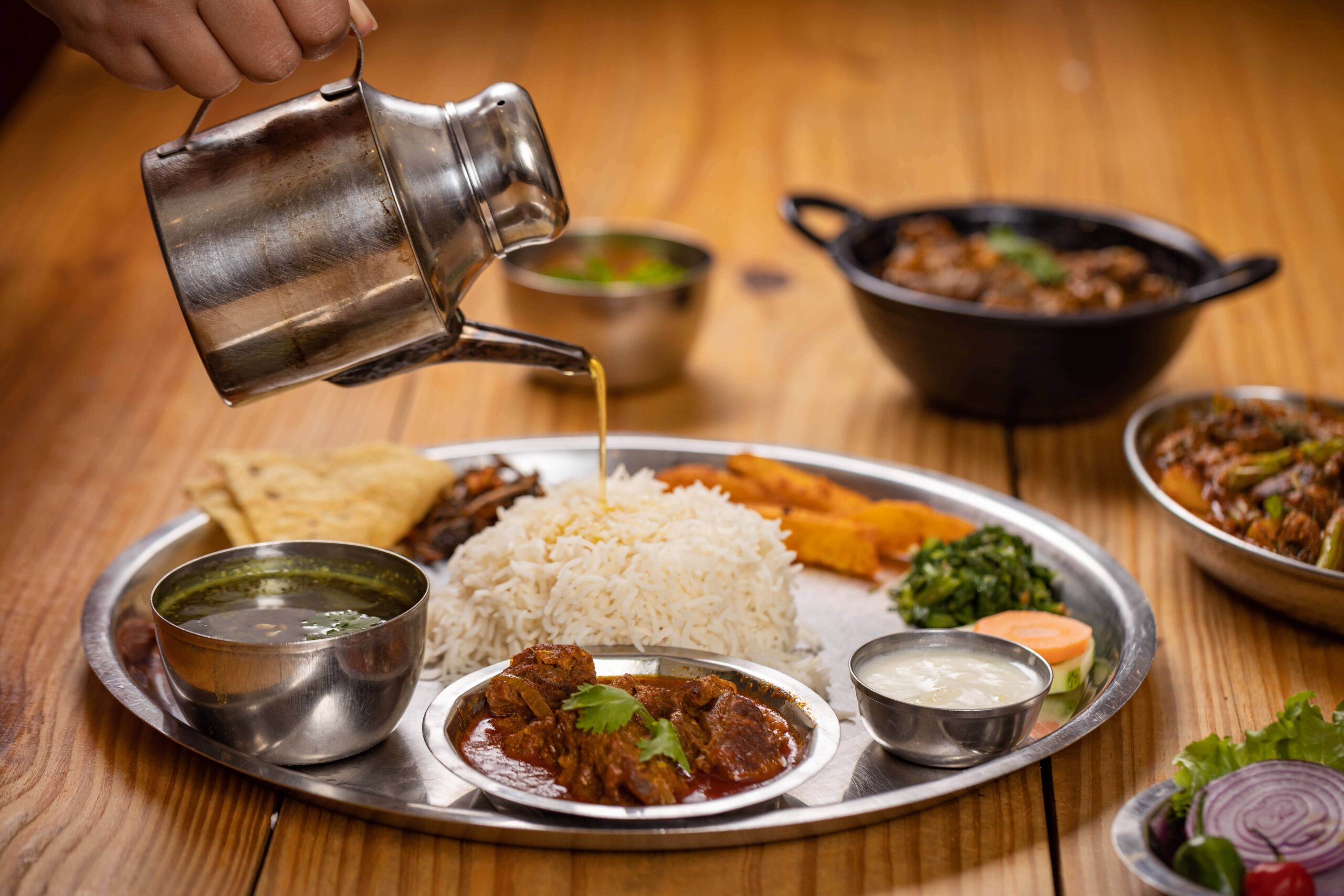A traditional Thakali Khana meal with rice, lentil soup, vegetables, pickles, and meat, served on a metal tray.