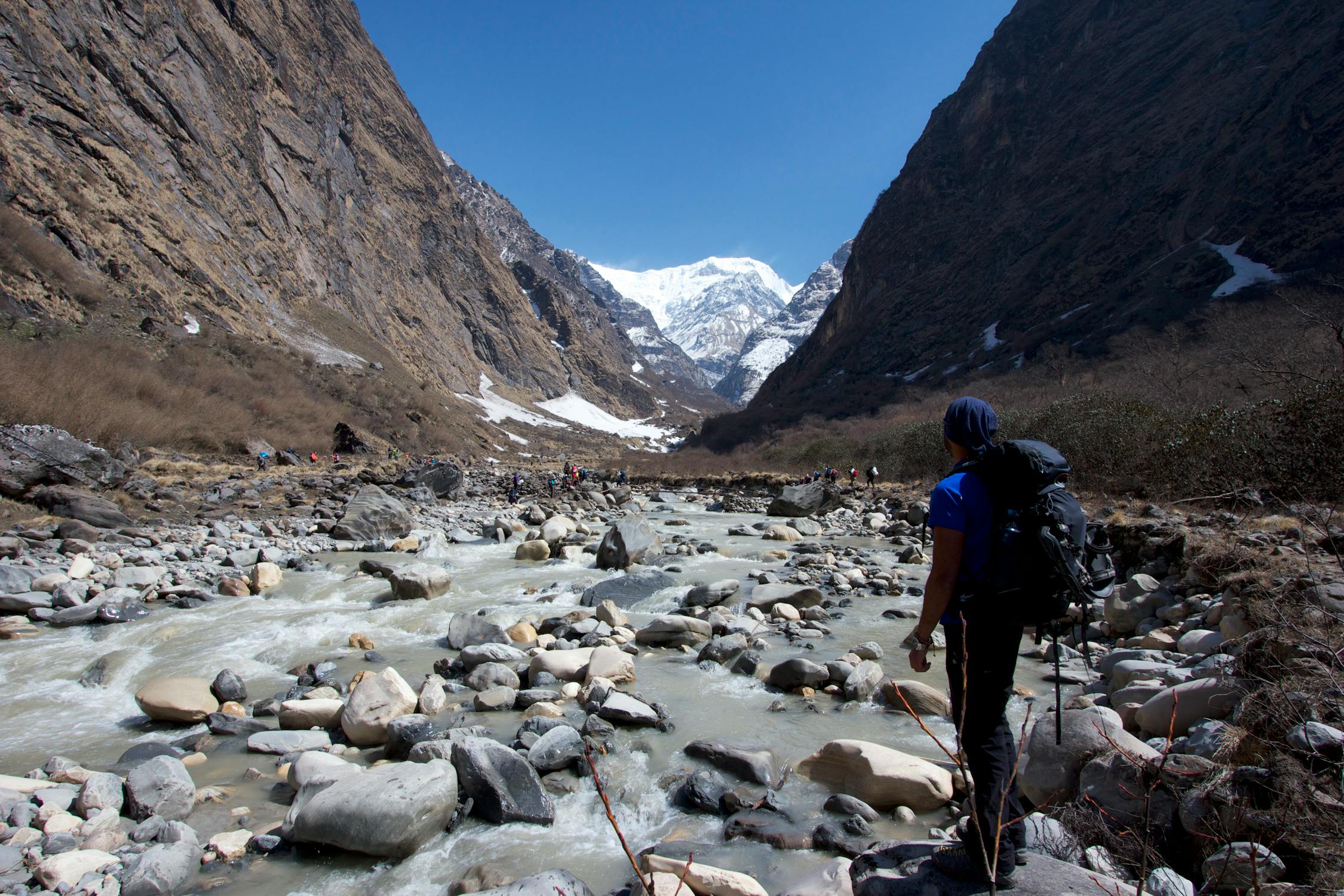 Trekker standing in the shallow waters of the Kali Gandaki River, looking at snow-covered Himalayan mountains.