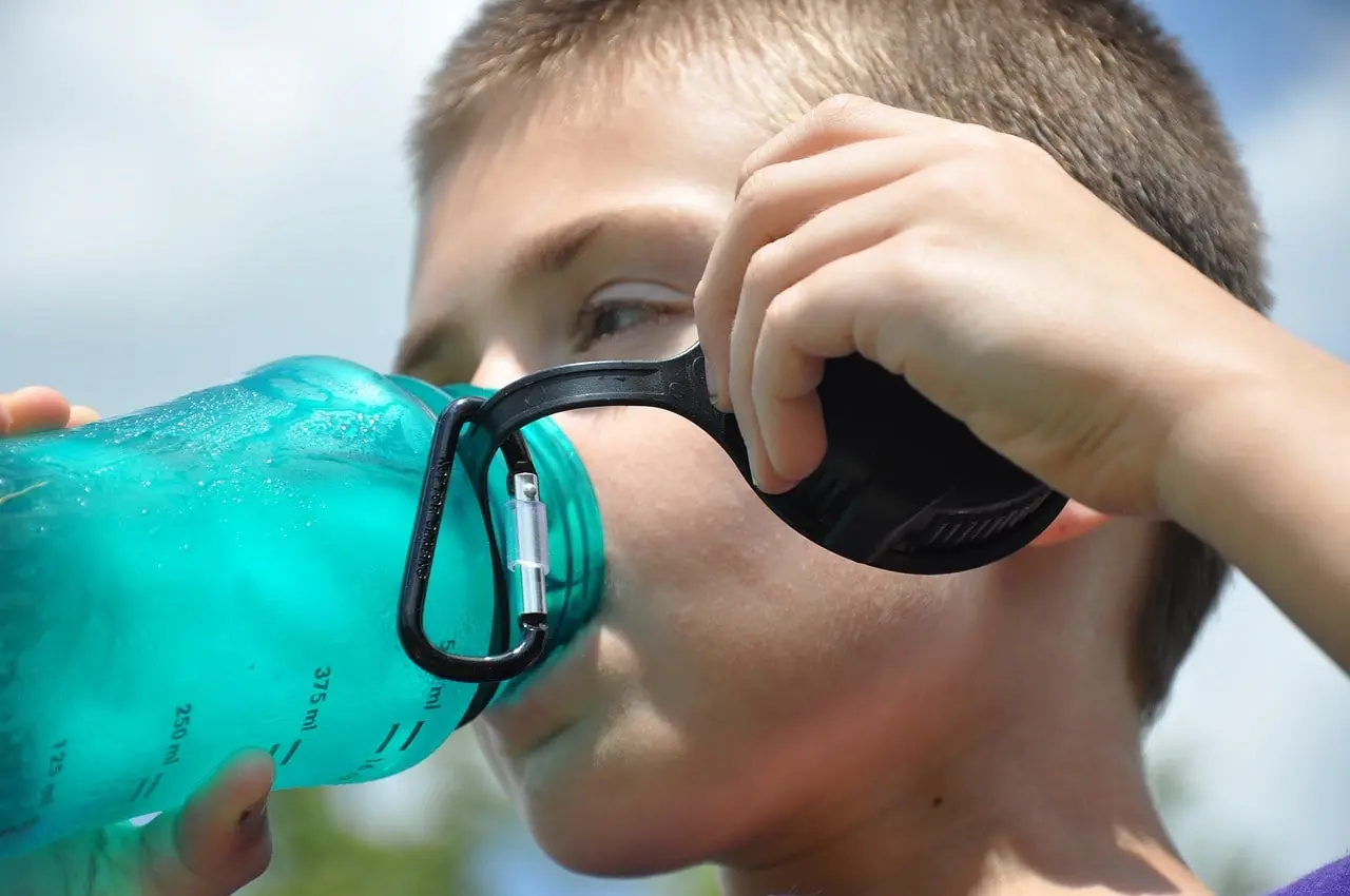 A young boy drinks water from a bottle while standing outside, showing the importance of hydration.